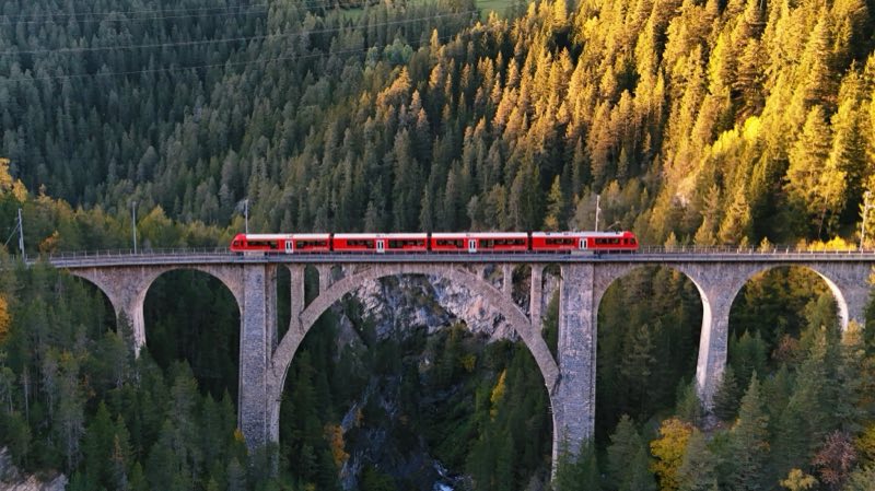 Red train on a stone viaduct with a large central arch over a gorge, lit by low evening sun.