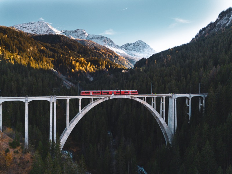Red train crossing a slender concrete arch viaduct over a forested valley, snowy peaks behind.