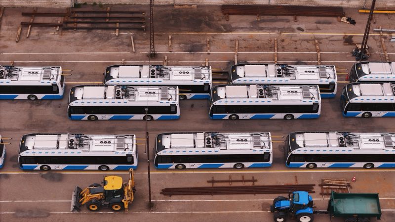 Blue and white trolleybuses lined up at a depot, a tractor and a digger in front.