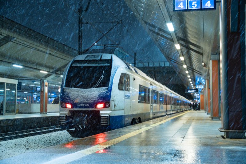 White and blue train standing at a covered platform at night in falling snow.