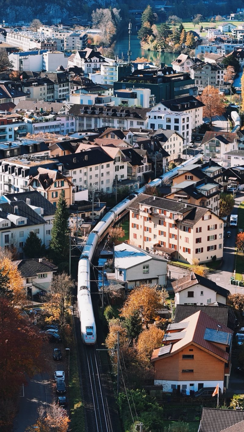 Long white train winding between apartment buildings and autumn trees in a town by a river.