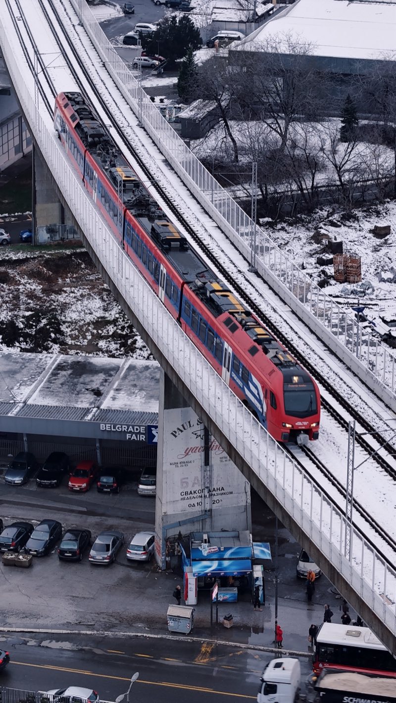 Red and blue train on an elevated railway curving over a snowy street.