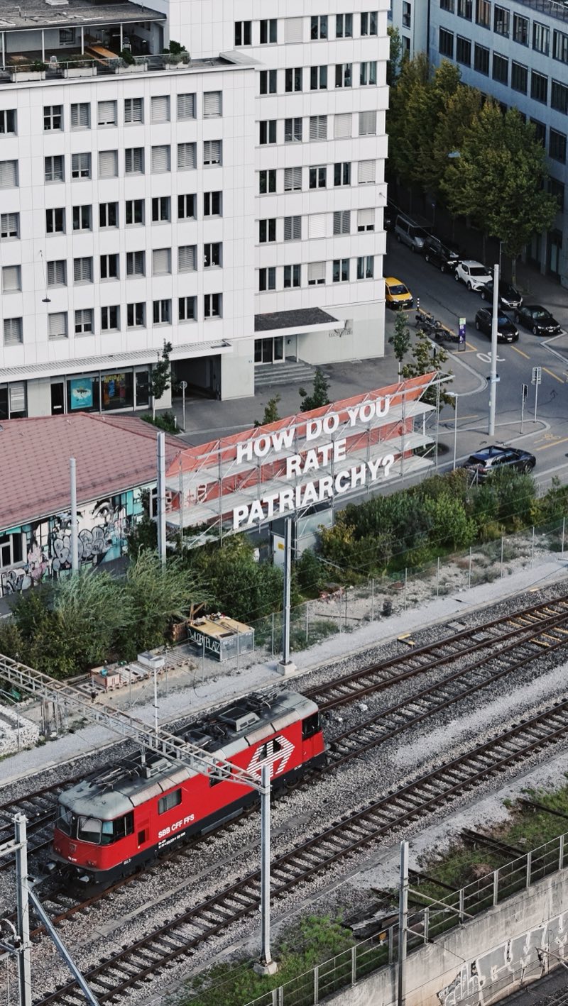 Red electric locomotive passing below an office building with a large sign.