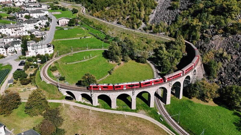 Red train on a spiral viaduct curving around a green meadow.