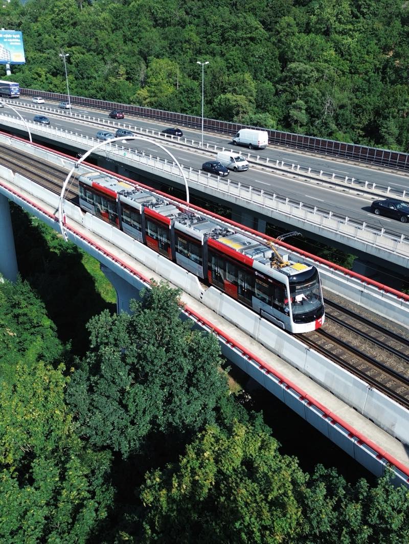 Red, white and grey tram on a viaduct beside a motorway, above dense trees.
