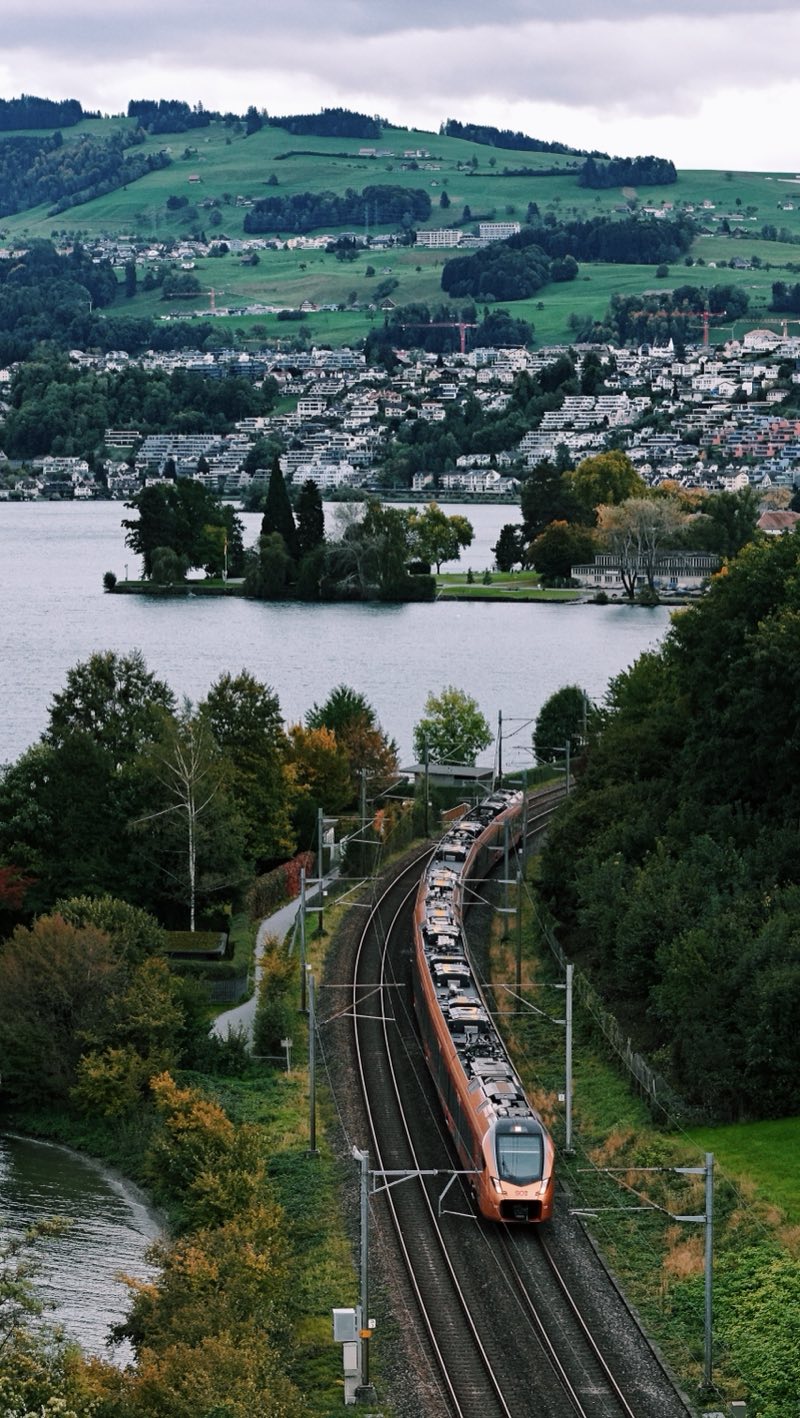 Red and white train on a curve beside a lake, a small island and a hillside town behind.