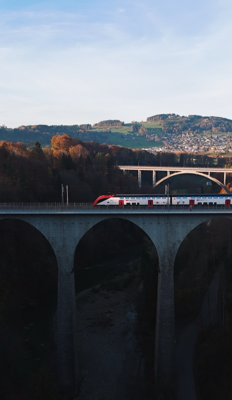 Red and white train on a dark viaduct, a second bridge and hills behind.