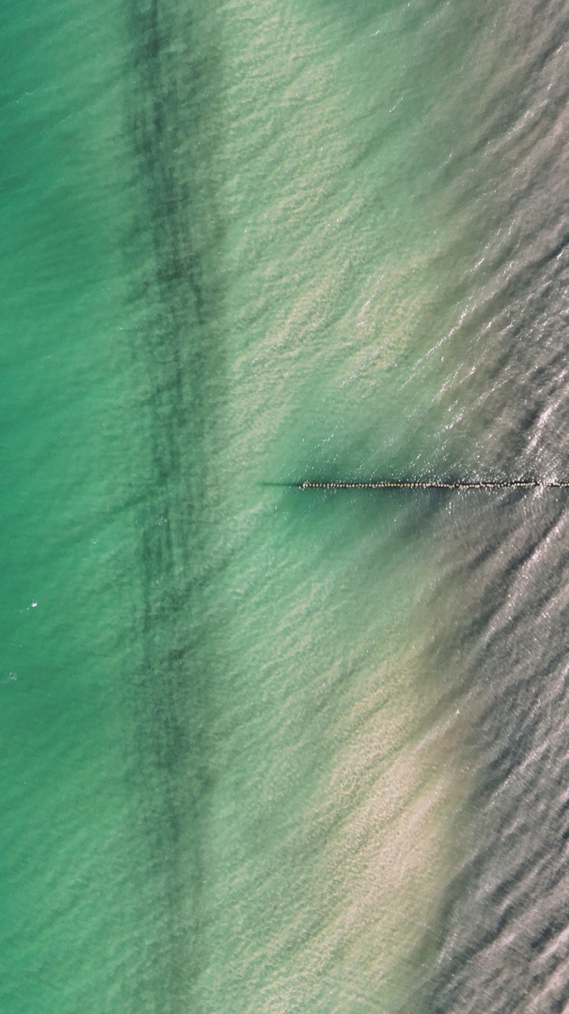 Shallow green sea seen from above, with a wooden groyne running out from the shore.