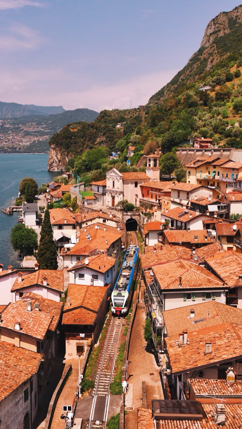 Train emerging from a tunnel between tiled roofs of a village on a steep lakeshore.