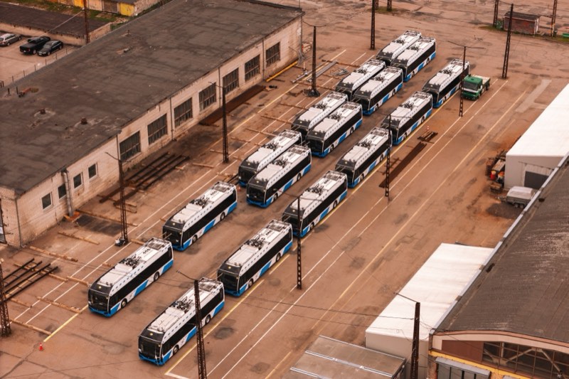 Two rows of blue and white trolleybuses parked in a depot yard next to an old brick building.