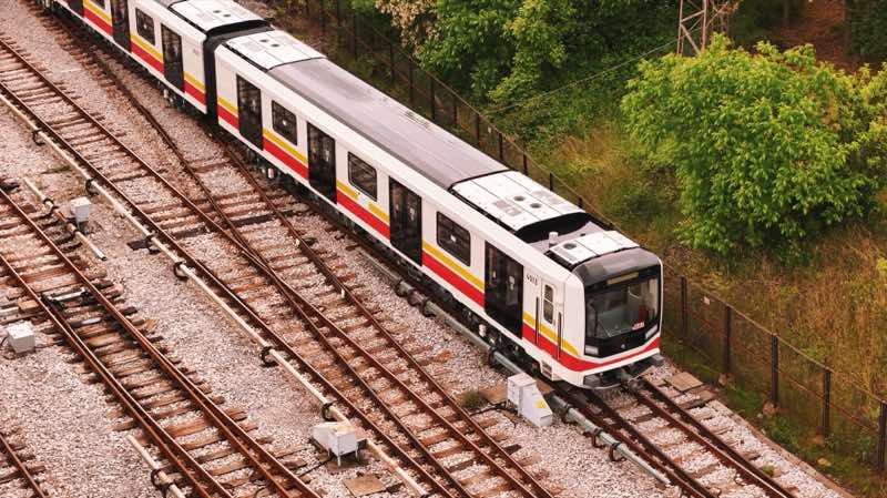 White metro train with red and yellow stripes on surface tracks next to trees.