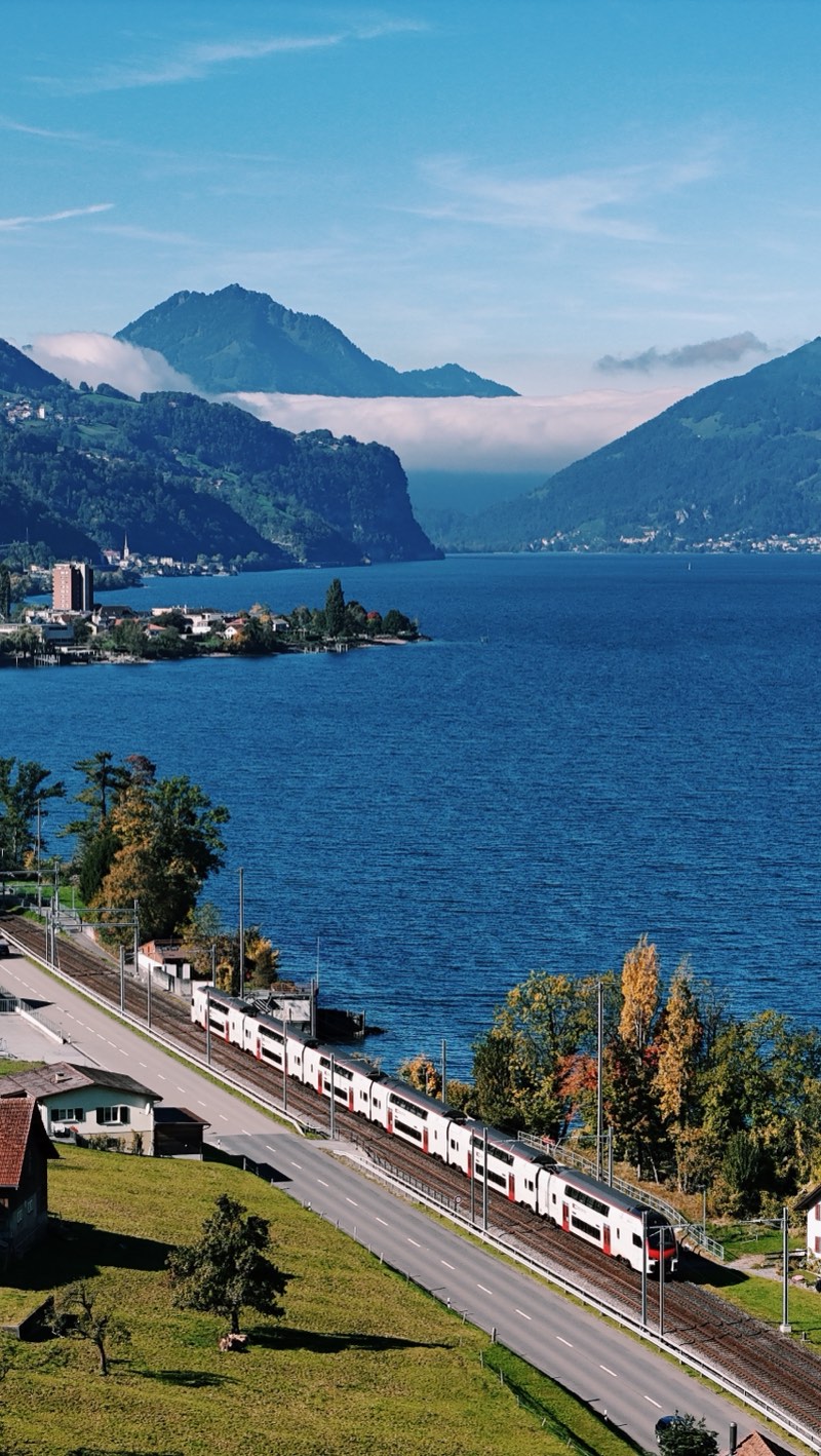 White and red double-deck train running along a deep blue lake, with mountains and low cloud behind.