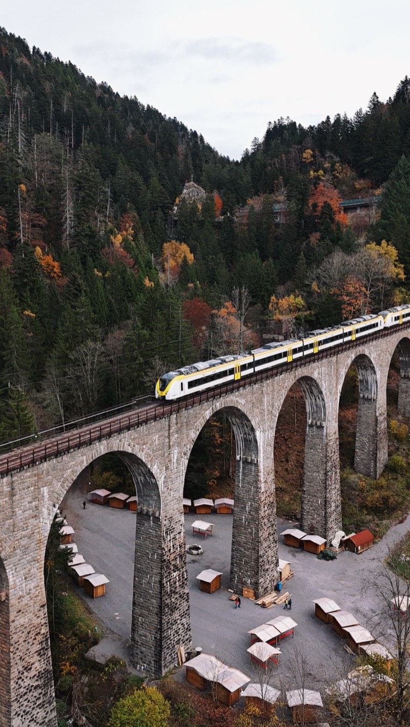 White and yellow train crossing a stone viaduct, stalls set out in the valley below.